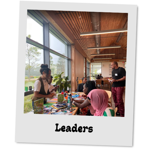 Photo styled as a tilted polaroid labeled "Leaders." In a bright, wood-paneled room with large windows, community members gather around a table spread with colorful art supplies, fabric, and books.